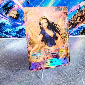 K&ouml;nnte beinhalten: Eine One Piece Sammelkarte mit der Figur Nico Robin mit langem dunklen Haar, bekleidet mit einem blauen Oberteil und einem rosa Rock. Die Karte zeigt den Text Nico Robin, Straw Hat Crew, 5000 power, 4 life sowie Spielregeln wie Banish und When Attacking.