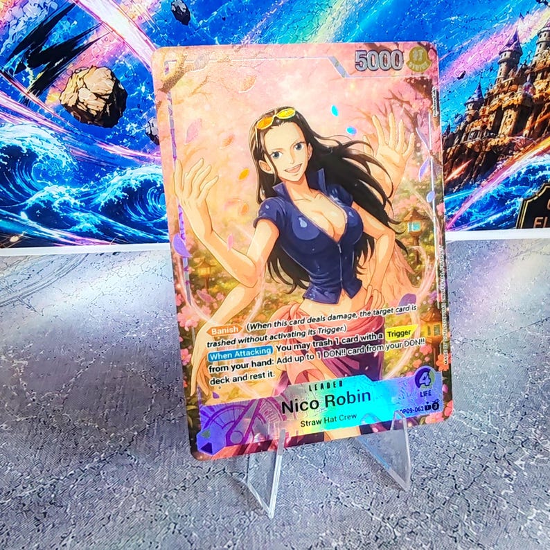 K&ouml;nnte beinhalten: Eine One Piece Sammelkarte mit der Figur Nico Robin mit langem dunklen Haar, bekleidet mit einem blauen Oberteil und einem rosa Rock. Die Karte zeigt den Text Nico Robin, Straw Hat Crew, 5000 power, 4 life sowie Spielregeln wie Banish und When Attacking.