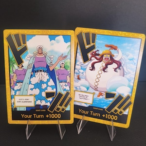 Puede incluir: Dos cartas coleccionables con bordes dorados e ilustraciones de personajes. Una carta presenta un personaje con un atuendo azul y blanco con el texto "GOD'S ARMY. GOD GUARDIANS." La otra muestra un personaje con sombrero amarillo y el texto "SATORI OF THE FOREST."