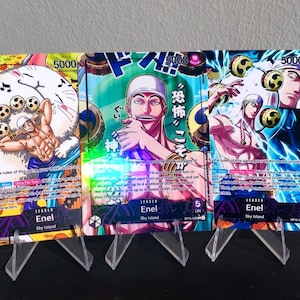 May include: Three collectible trading cards featuring the character Enel from the "One Piece" anime series. Each card displays a different illustration of Enel, with Japanese text and card details. The cards are displayed on clear plastic stands.