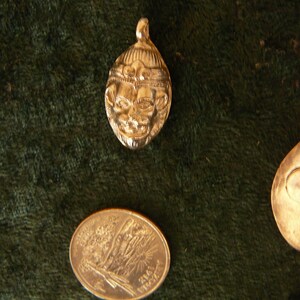 May include: Two gold-colored pendants on a green velvet background. One pendant features a detailed face with a crown, the other is a smooth oval shape.