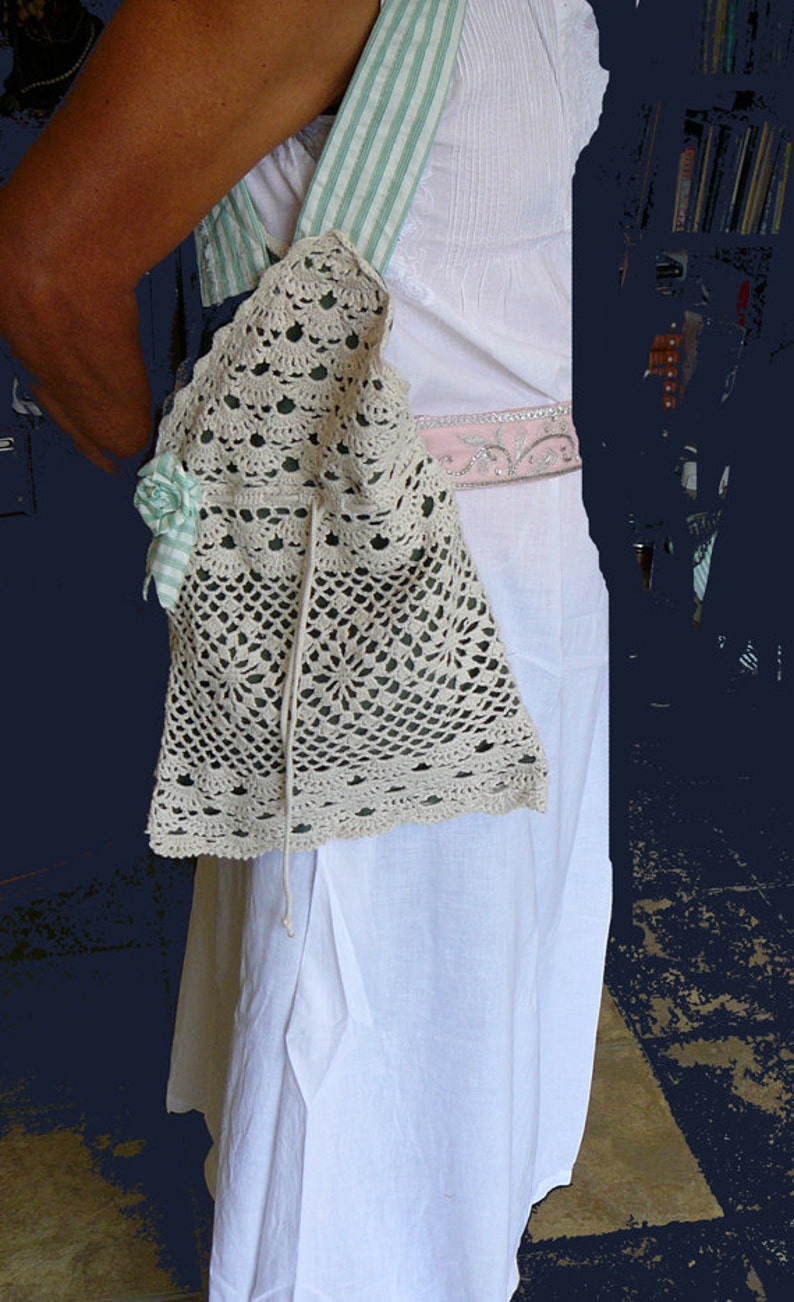 May include: A white crocheted bag with a green and white striped strap. The bag has a floral embellishment and a drawstring closure.