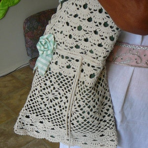 May include: A white crocheted bag with a green and white ribbon flower accent. The bag has a drawstring closure and is worn over the shoulder.