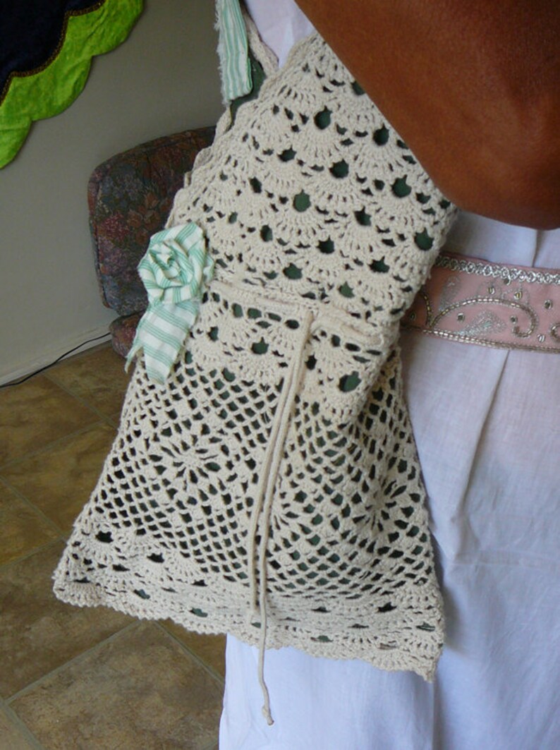 May include: A white crocheted bag with a green and white ribbon flower accent. The bag has a drawstring closure and is worn over the shoulder.