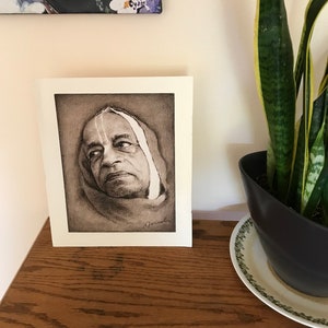 May include: A framed pencil portrait of a man with a white head covering. The artwork is displayed on a wooden surface, with a potted snake plant and a painting of flowers in the background.