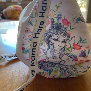 May include: A white fabric bag featuring a detailed illustration of a figure with curly hair playing a flute, surrounded by colorful floral and peacock feather motifs. The shoulder strap is printed with the text Rama Hare Hare.