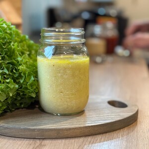 Puede incluir: Un tarro de cristal transparente lleno de un líquido amarillo-verdoso, posiblemente aderezo para ensaladas, sobre una tabla de cortar de madera. Un gran manojo de lechuga verde está a la izquierda del tarro. La tabla tiene un recorte en forma de asa.