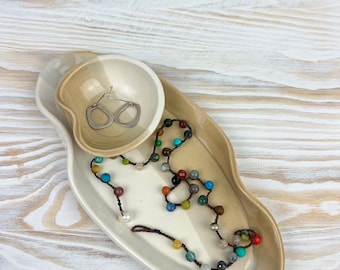 May include: A ceramic jewelry tray with a small bowl and a larger, oblong section. The tray is two-toned, with a light beige and brown glaze. A beaded necklace with various colored beads and a pair of silver earrings rest on the tray.