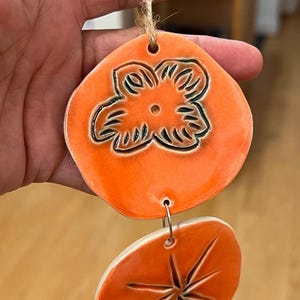 May include: Orange ceramic wind chime with a flower design on the top piece and a starburst design on the bottom piece. The pieces are connected with metal rings and a natural twine string.