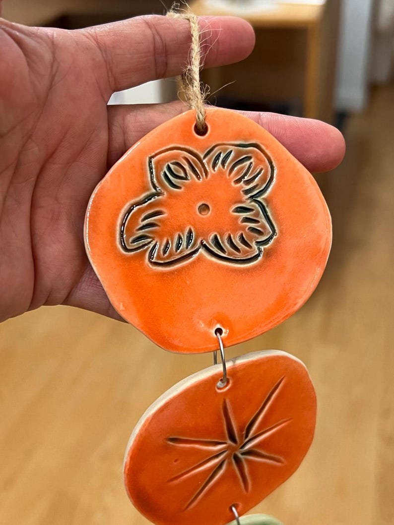 May include: Orange ceramic wind chime with a flower design on the top piece and a starburst design on the bottom piece. The pieces are connected with metal rings and a natural twine string.