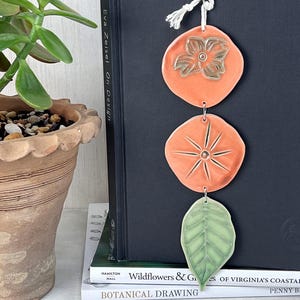 May include: A ceramic wall hanging with three orange and green shapes. The top shape is a flower, the middle is a starburst, and the bottom is a leaf. The wall hanging is hanging on a black book. The text "Eva Zeisel On Design" is visible.