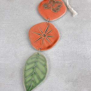 May include: A ceramic wind chime featuring three decorative elements: a peach-colored flower-shaped piece, a peach-colored starburst-shaped piece, and a green leaf-shaped piece. The pieces are connected with metal rings and a white string.