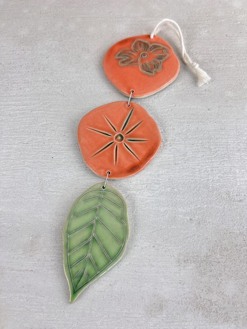 May include: A ceramic wind chime featuring three decorative elements: a peach-colored flower-shaped piece, a peach-colored starburst-shaped piece, and a green leaf-shaped piece. The pieces are connected with metal rings and a white string.