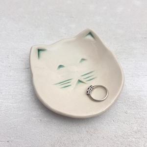 May include: A cream-colored ceramic ring dish shaped like a cat's face. The dish has green accents for the eyes and whiskers. A silver ring with three small stones rests inside the dish. The background is a light gray surface.