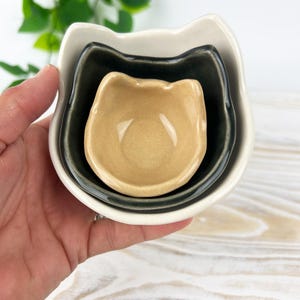 May include: A set of three ceramic cat-shaped nesting bowls in cream, dark gray, and tan. The bowls are stacked inside each other, with the smallest bowl in the center. The bowls are held in a hand.
