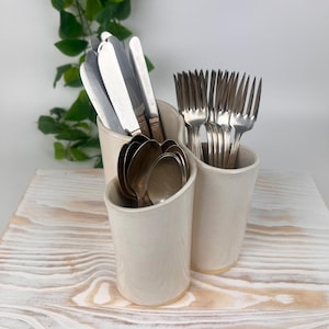 May include: A cream-colored ceramic utensil holder with three compartments, filled with silverware. The silverware includes knives, forks, and spoons. The holder sits on a light-colored wooden surface, with green foliage in the background.