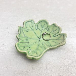 May include: A light green ceramic leaf-shaped ring dish with a gold ring. The dish has detailed leaf vein patterns and a glossy finish. The ring has three small stones. The dish is on a light gray surface.