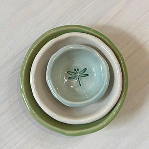 May include: A set of three ceramic bowls in varying sizes and colors. The largest bowl is a light green, the middle bowl is white, and the smallest bowl is a light blue with a dragonfly design.
