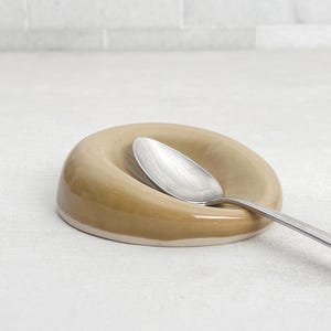 May include: A ceramic spoon rest in a light brown color with a white base. A silver spoon rests inside the curved indentation. The spoon rest has a smooth, glossy finish and is designed to hold a spoon.