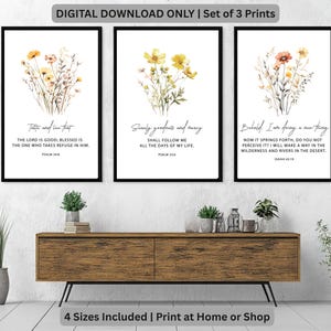 Peut inclure: Ensemble de trois impressions d'art encadrées avec des arrangements floraux à l'aquarelle en jaune, orange et beige. Chaque impression a un cadre noir et comprend un texte inspirant de la Bible. Les impressions sont affichées au-dessus d'un meuble en bois.