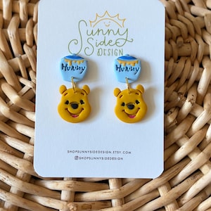 May include: A pair of dangle earrings featuring a yellow bear face charm and a light blue honey pot charm with the word Hunny written on it. The earrings are displayed on a white card with the text Sunny Side Design and social media handles.