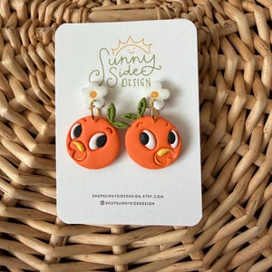 May include: A pair of orange, round earrings with cartoon-like orange bird faces, white daisy accents, and green leaves. The earrings are displayed on a white card with the text "Sunny Side Design" and shop information.