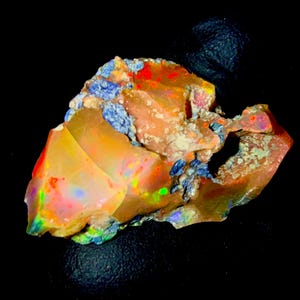 Peut inclure: Un spécimen d'opale brut et non poli présentant une base orange et jaune translucide avec des éclats vibrants de rouge, de vert et de bleu. La pierre a une forme irrégulière et dentelée avec des taches d'inclusions minérales gris-bleu.