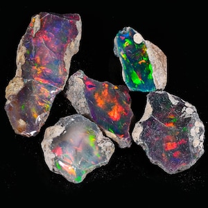 Puede incluir: Cinco piedras de ópalo en bruto con colores vibrantes como rojo, verde, azul y naranja. Los ópalos tienen una textura rugosa y natural sobre un fondo negro. Estas piedras preciosas son ideales para la creación de joyas o coleccionar.