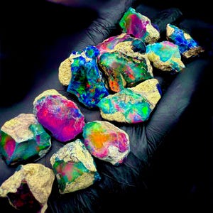 May include: A collection of rough opal gemstones, showcasing vibrant colors including blues, greens, pinks, and oranges. The opals have a rough, natural texture with a beige matrix. The gemstones are displayed against a black background.