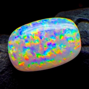 May include: A polished, oval-shaped opal gemstone featuring a vibrant play-of-color with bright flashes of neon green, electric blue, hot pink, and golden yellow against a translucent, milky white base, set against a dark, textured background.