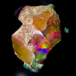 Könnte beinhalten: Ein roher Opal-Edelstein mit einer rauen, unregelmäßigen Form. Der Stein zeigt eine Mischung aus Farben, darunter Orange, Gelb und Rosa, mit leuchtenden Blitzen von Rot, Grün und Blau. Der Opal ist vor einem schwarzen Hintergrund platziert.