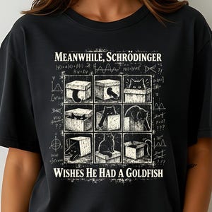 Puede incluir: Una camiseta negra de manga corta con un estampado gráfico blanco de nueve cajas que contienen gatos. El texto dice: MEANWHILE, SCHRÖDINGER WISHES HE HAD A GOLDFISH. El diseño incluye fórmulas matemáticas y símbolos científicos de fondo.