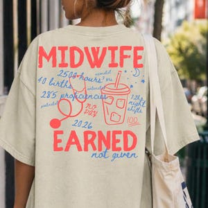 May include: A beige t-shirt with the word "MIDWIFE" in red block letters. Below, the shirt features a graphic of a stethoscope, a drink, and text that reads "EARNED not given". The shirt is part of a tote bag.