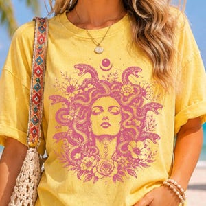 Peut inclure: Un t-shirt jaune à manches courtes avec une impression graphique magenta représentant un visage entouré de serpents et de fleurs. Le vêtement présente une encolure ronde et une coupe décontractée. Une bandoulière à motifs repose sur l'épaule gauche.
