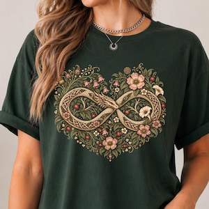 以下が含まれることがあります： ダークフォレストグリーンのTシャツで、中央にハート型のインフィニティシンボルのグラフィックが描かれています。デザインには、精巧な鱗を持つとぐろを巻いたドラゴンが組み込まれており、ピンクの花や緑の葉などの植物の要素で囲まれています。