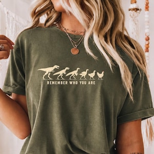 Op de afbeelding: Een olijfgroen T-shirt met korte mouwen en een afbeelding van zes wezens die evolueren van een dinosaurus naar een kip. Onder de afbeelding staat de tekst REMEMBER WHO YOU ARE in een eenvoudig, lichtgekleurd, schreefloos lettertype.