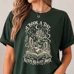以下が含まれることがあります： 暗い緑色の半袖Tシャツ。開いた本から丘の上の城へと続く道が描かれたクリーム色のグラフィックプリントが施されています。星、雲、木々に囲まれて「A BOOK A DAY KEEPS REALITY AWAY」というテキストが記されています。