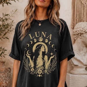 May include: A black short-sleeved t-shirt featuring a gold graphic print of a woman with long hair surrounded by a moon phase cycle, floral elements, and the word LUNA. The design includes a star and a circular halo behind the figure.