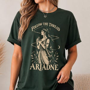 Peut inclure: Un t-shirt à manches courtes vert foncé avec une impression graphique représentant une femme en robe classique tenant un récipient. Le texte "FOLLOW THE THREAD" est arqué au-dessus de la figure et le nom "ARIADNE" est imprimé en dessous.