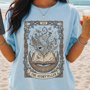 May include: Pale blue t-shirt with a detailed graphic that reads "THE STORYTELLER". The design features an open book, a crescent moon, and botanical elements within a decorative frame. The shirt has short sleeves.