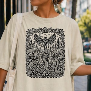 Puede incluir: Una camiseta de color crema con un estampado gráfico detallado en tinta negra. El diseño muestra una figura central de Bigfoot rodeada por una escena forestal con un búho, un ciervo, una serpiente, una ardilla, un zorro, un conejo y setas.