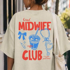 May include: Beige t-shirt featuring a red and blue graphic on the back. The graphic design includes the words "tired MIDWIFE CLUB" and illustrations of a bow, coffee cup, syringe, and stethoscope. The text "i'm fine just tired" is also included.