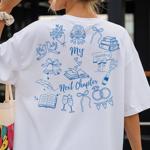 May include: White t-shirt featuring blue wedding-themed illustrations, such as a car, doves, books, and rings. The text "my Next Chapter" is printed on the back. The shirt has short sleeves and a loose fit, suitable for casual wear.
