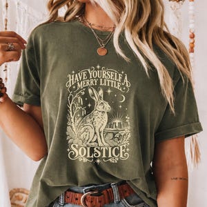 以下が含まれることがあります： ウサギ、星、そして「HAVE YOURSELF A MERRY LITTLE SOLSTICE」という文字がクリーム色でプリントされた、オリーブグリーンの半袖Tシャツ。クルーネックで、茶色のレザーベルトとブルーのデニムジーンズを合わせて着用しています。