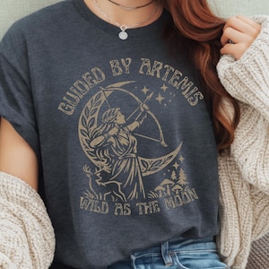 Peut inclure: T-shirt gris foncé avec une illustration beige d'Artémis, déesse grecque de la chasse, avec un arc et des flèches, sur un croissant de lune. Le texte dit "GUIDED BY ARTEMIS" et "WILD AS THE MOON".