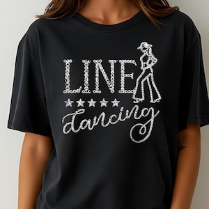 Könnte beinhalten: Schwarzes T-Shirt mit dem Schriftzug "LINE DANCING" in einem glitzernden, strassartigen Schriftstil. Das Design beinhaltet eine Silhouette einer Person beim Line Dance und fünf Sterne. Ein goldenes Armband ist sichtbar.
