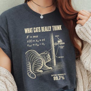 Può includere: Una t-shirt grigio antracite scuro con una stampa grafica intitolata WHAT CATS REALLY THINK. Il design include equazioni fisiche, un diagramma di un gatto che insegue un topo dietro un palo e il testo CAPTURE PROBABILITY: 99.7% in color crema.