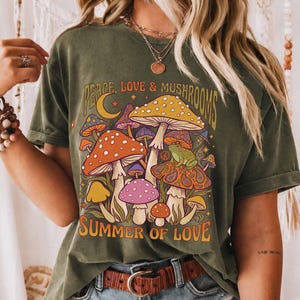 Op de afbeelding: Olijfgroen T-shirt met een kleurrijke afbeelding van paddenstoelen, een kikker en de tekst "Peace, Love & Mushrooms" en "Summer of Love". Het ontwerp bevat oranje, gele en paarse paddenstoelen. Het shirt heeft korte mouwen en een relaxte pasvorm.