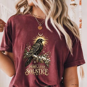 Puede incluir: Una camiseta de manga corta color granate con un gráfico de un cuervo negro posado en una rama de pino nevada con piñas. Sobre el ave hay un sol dorado y una luna creciente. El texto dice, Have yourself a merry little solstice.