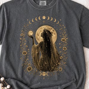 May include: Dark gray t-shirt featuring a golden illustration of a woman with long hair, a floral crown, and a flowing dress, holding a moon. The design includes a large moon, moon phases, and floral accents.